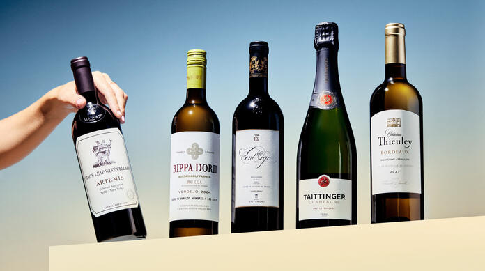 Various wine bottles lined up against blue background with person's hand grabbing the bottle on the end.