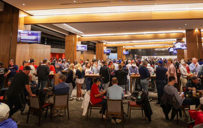 A crowded indoor stadium lounge with fans standing and sitting at tables beneath wood-paneled ceilings and multiple TV screens.