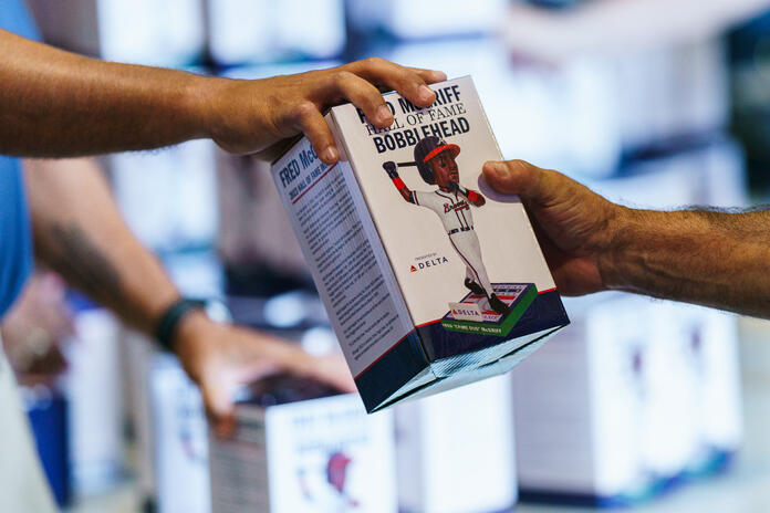 Two hands exchange a boxed “Hall of Fame Bobblehead,” showing a baseball player figurine on the packaging, with stacks of similar boxes in the background.