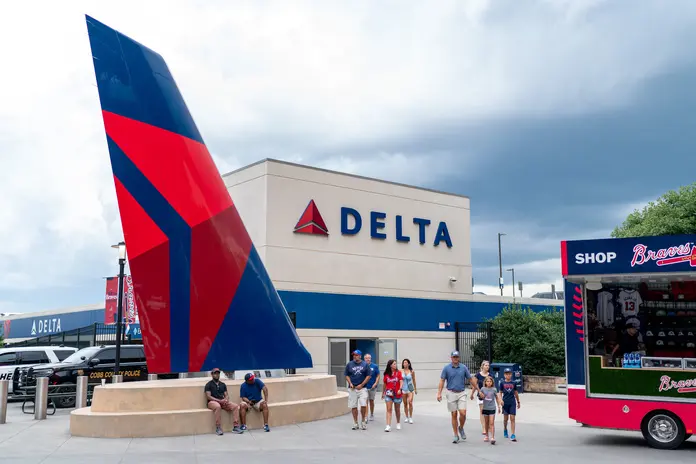 People walk outside a stadium concourse near a large Delta airplane tail sculpture and a Delta-branded building, with a Braves merchandise shop nearby.