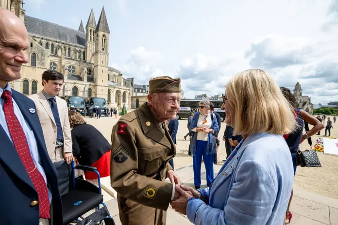 A uniformed World War II–era veteran shakes hands with an attendee in front of a historic stone church, with a small group of people gathered nearby at an outdoor commemorative event.