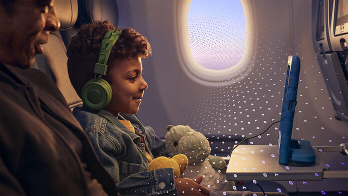  A child seated on an airplane uses a tablet while wearing headphones and holding a stuffed toy, with another seated adult and an airplane window visible, overlaid with a subtle digital pattern.