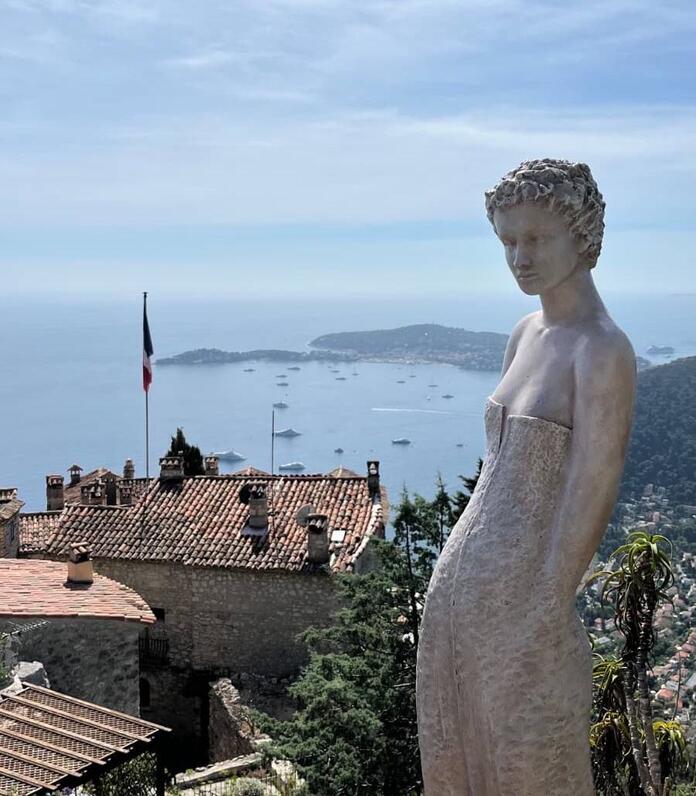 Stone statue overlooking a Mediterranean bay with terracotta rooftops and anchored boats below.