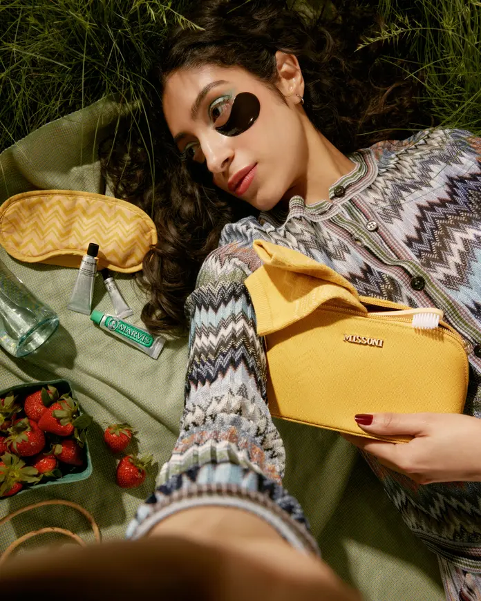 Person lying on grass with a yellow Missoni pouch, eye mask, skincare items, toothpaste, and strawberries arranged on a picnic cloth.