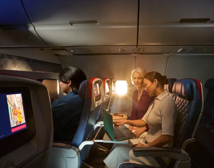 Two female Delta customers on board an aircraft working on laptops, with warm sunlight streaming through the windows.