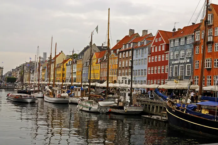 Colorful waterfront buildings line a canal with docked boats and masts, reflecting on the water under an overcast sky.