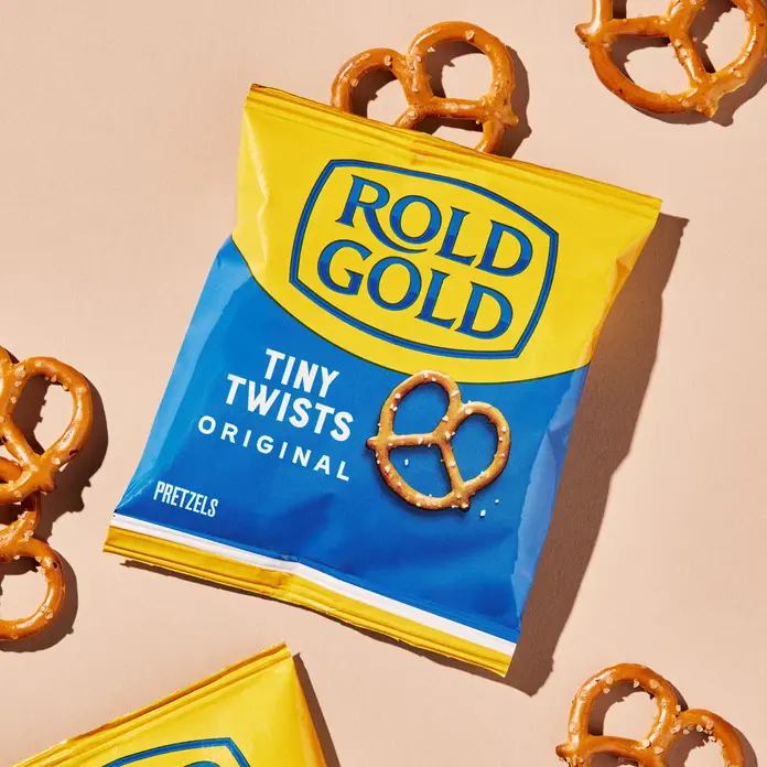 Close-up of a yellow and blue Rold Gold Tiny Twists Original pretzel bag surrounded by scattered mini pretzels on a light background.
