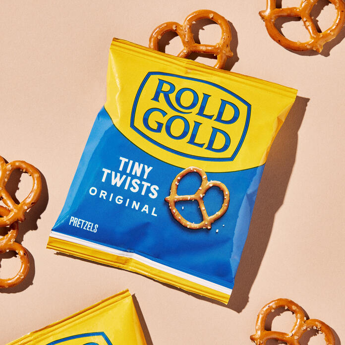 Close-up of a yellow and blue Rold Gold Tiny Twists Original pretzel bag surrounded by scattered mini pretzels on a light background.