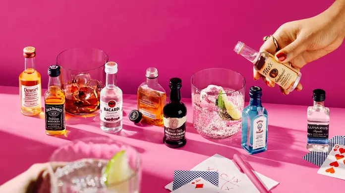 Mini bottles of liquor and cocktails arranged on a bright pink surface, with a hand pouring vodka into a glass with ice and lime, surrounded by playing cards and bar accessories.