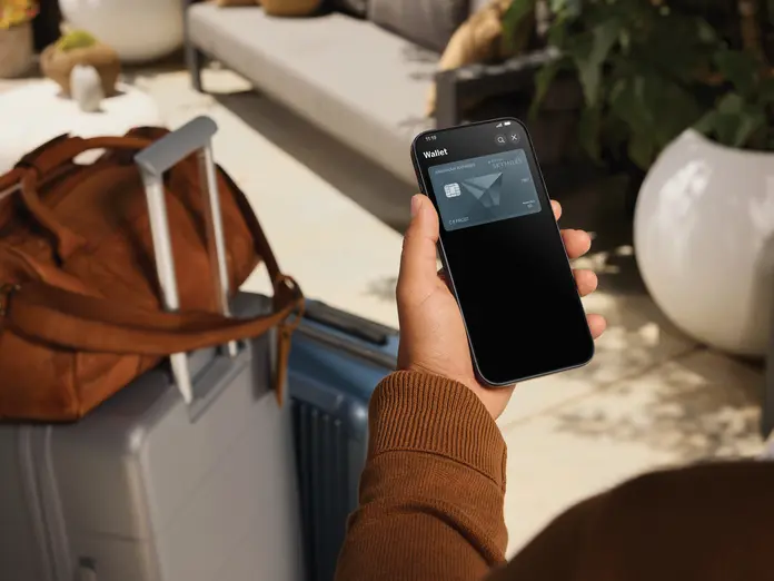 Person holding a smartphone displaying a Delta SkyMiles American Express Card in a digital wallet, with a suitcase and travel bag nearby in a lounge setting.