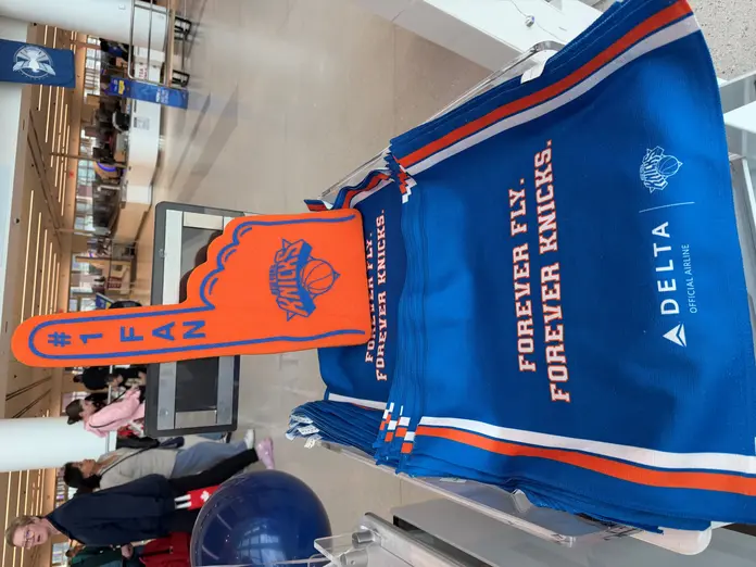 Bright orange New York Knicks foam finger displayed on a stand of blue Delta‑branded rally towels reading “Forever Fly. Forever Knicks” in an airport terminal, with travelers and security area in the background.