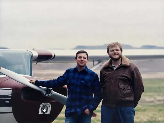 Two individuals stand beside a small propeller airplane on a grassy airfield, with one resting a hand on the wing and mountains visible in the background.