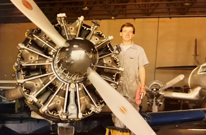 Person in a maintenance jumpsuit standing beside a vintage radial aircraft engine with a large propeller in a hangar.