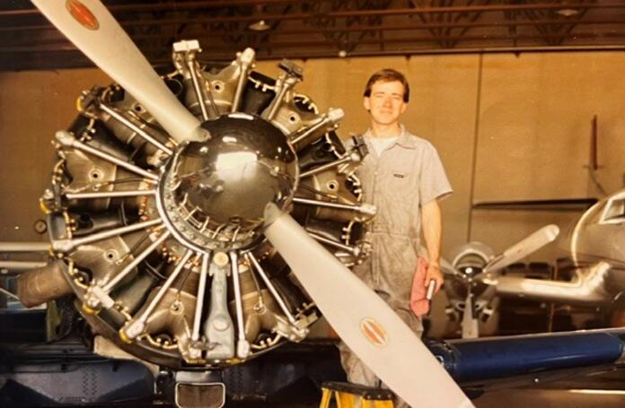 Person in a maintenance jumpsuit standing beside a vintage radial aircraft engine with a large propeller in a hangar.