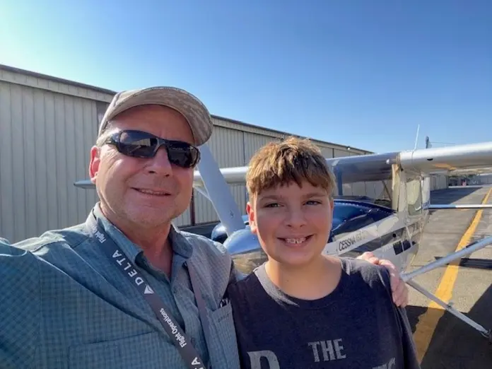 Adult wearing sunglasses and a cap stands beside a child, both smiling next to a small airplane on a sunny airfield.