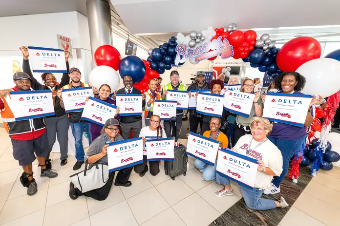 Group of Delta employees and guests pose beneath a Braves-themed balloon arch, holding commemorative Delta and Braves signs during the Gate A25 celebration.