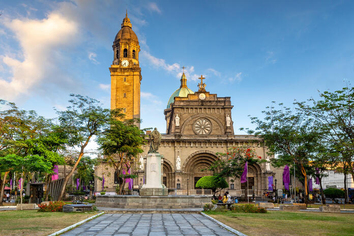 Historic cathedral with a tall clock tower, framed by trees and a central monument in a landscaped plaza under a blue sky.