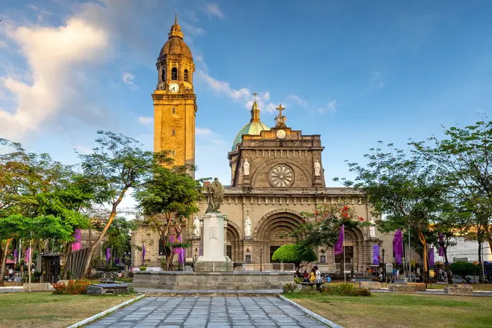 Historic cathedral with a tall clock tower, framed by trees and a central monument in a landscaped plaza under a blue sky.