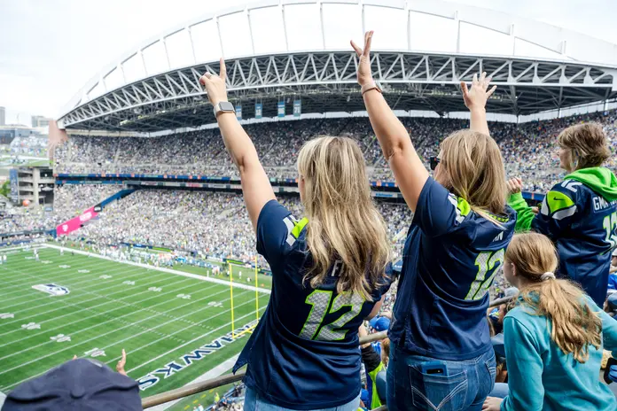 Fans wearing Seattle football jerseys raise their arms while watching a game from the stands of a crowded stadium.