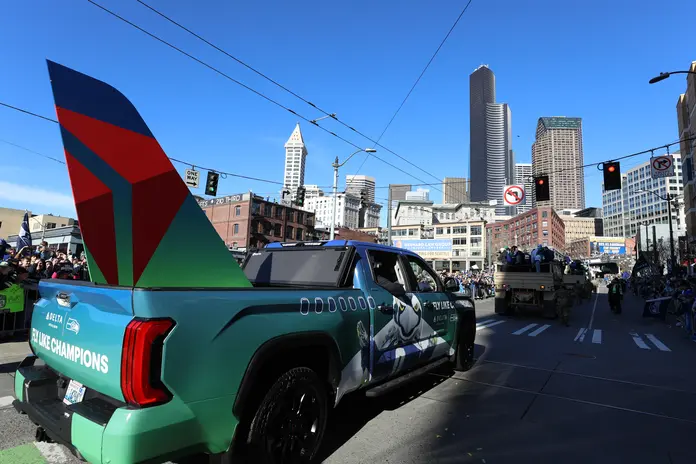 Delta-branded vehicle carrying a large airplane tail design during a parade through downtown Seattle, with crowds lining the streets and the city skyline in the background.