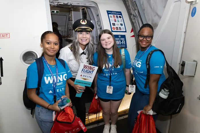 Students wearing WING Flight shirts pose with a pilot at an aircraft doorway during a Girls in Aviation Day experience.