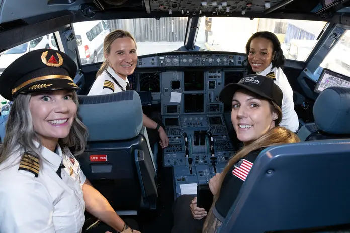 Aviation professionals pose in an aircraft cockpit, seated at the flight controls and pilot seats, during a Girls in Aviation Day event.