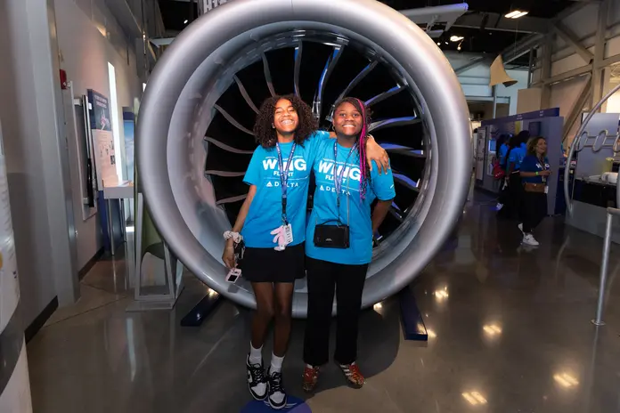 Two students wearing WING Flight shirts pose in front of a large aircraft engine display during a visit to the Airbus facility.