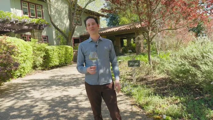 Person holding a glass of white wine on a garden path outside a vineyard-style building surrounded by trees and landscaping.