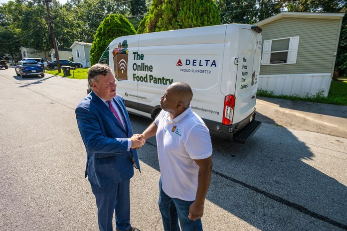Two people shake hands beside a Delta-supported Online Food Pantry delivery van in a residential neighborhood.