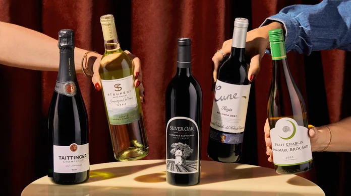 Five wine bottles on a table, with hands reaching in to hold and select different bottles against a dark red curtain backdrop.