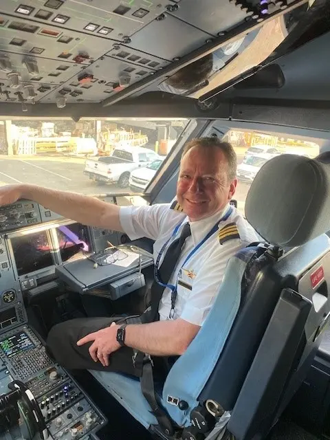 Person in a pilot uniform seated in an aircraft cockpit, smiling with controls and instruments visible.