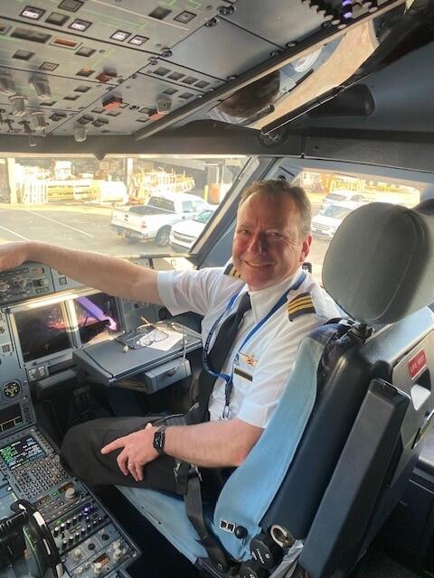 Person in a pilot uniform seated in an aircraft cockpit, smiling with controls and instruments visible.