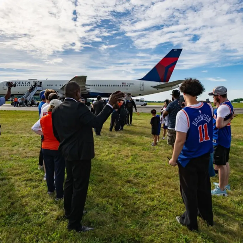 Spectators stand on grass watching a Delta airplane taxi on the runway at an airport.