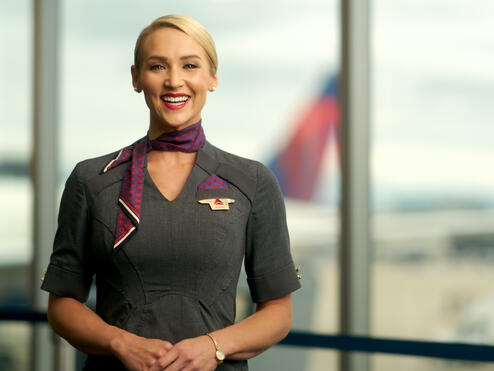 Delta flight attendant smiling, wearing gray dress and purple neck scarf.