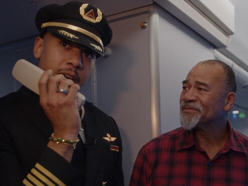 Airline pilot in uniform stands inside an aircraft cabin near the galley, holding an intercom handset. Another person wearing a red plaid shirt is standing beside the pilot.