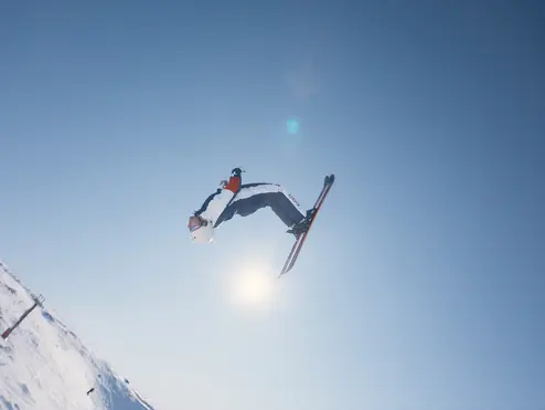 A skier performs a high aerial trick against a clear blue sky on a snowy mountain slope