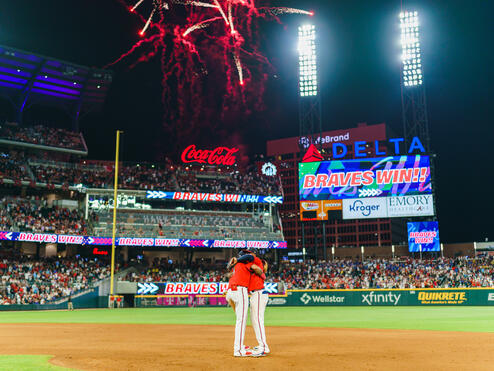 Fireworks light up a baseball stadium at night as two players stand together on the infield, with a scoreboard displaying “Braves Win” and a packed crowd in the background.