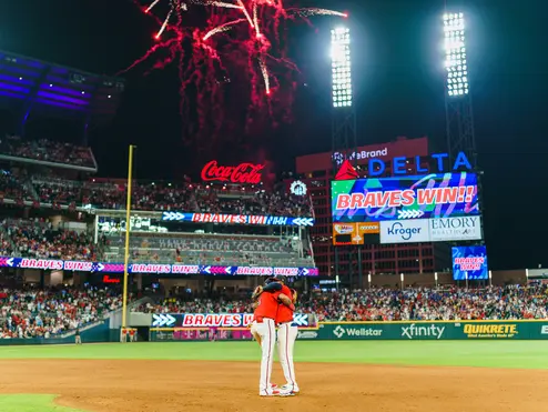 Fireworks light up a baseball stadium at night as two players stand together on the infield, with a scoreboard displaying “Braves Win” and a packed crowd in the background.