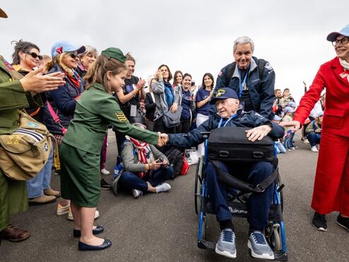 A seated veteran in a wheelchair shakes hands with a child in a green uniform as a crowd lines the roadway, applauding during a commemorative event.
