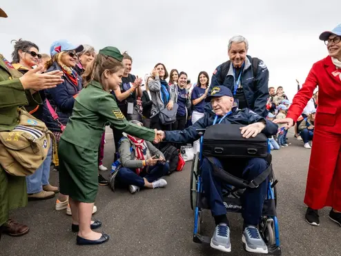 A seated veteran in a wheelchair shakes hands with a child in a green uniform as a crowd lines the roadway, applauding during a commemorative event.