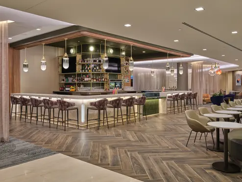 Lounge bar with high‑back stools, illuminated shelving, warm lighting and adjacent seating areas with small tables