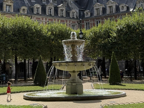 Stone fountain in a leafy Paris square, framed by manicured trees and historic red-brick buildings with slate roofs.
