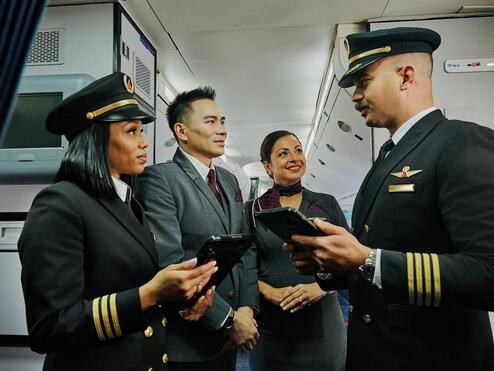 Two Delta pilots and two Delta flight attendants stand together in an airplane aisle, holding tablets and talking.