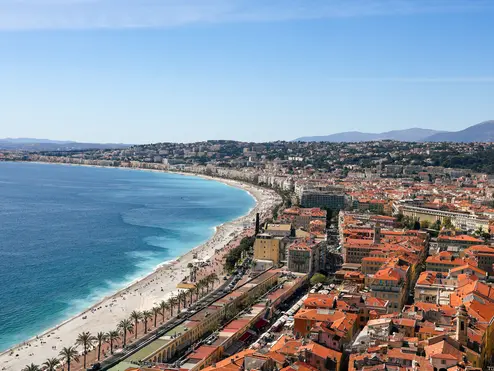 Aerial view of a curved Mediterranean beach and waterfront city with red-tiled roofs.