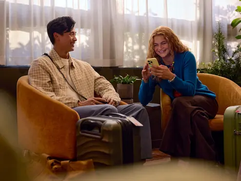 Two travelers sit in lounge chairs with rolling suitcases nearby, one looking at a smartphone, in a softly lit indoor waiting area with plants and sheer curtains.