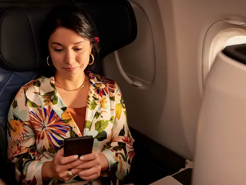 Passenger seated by an airplane window holds a smartphone while sitting in a premium cabin seat, with soft lighting and aircraft interior visible.