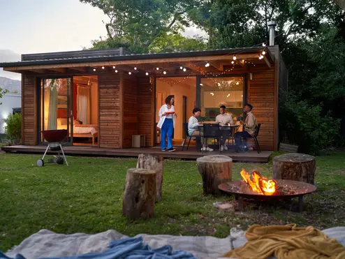 People dining outside a modern wooden cabin at dusk, with string lights, a fire pit and trees surrounding the yard.