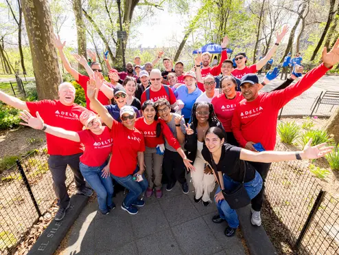 A large group of Delta volunteers wearing red “Delta in the Community” shirts stand together in a park with arms raised, gathered along a paved path surrounded by trees.