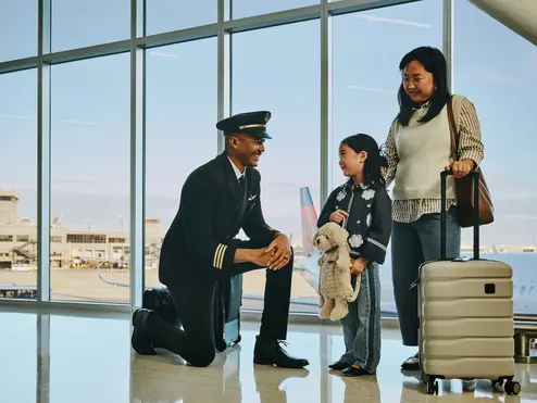 Delta pilot kneeling beside a young child holding a stuffed animal at an airport gate, while an adult stands nearby with rolling luggage; airplane visible through large windows in the background.