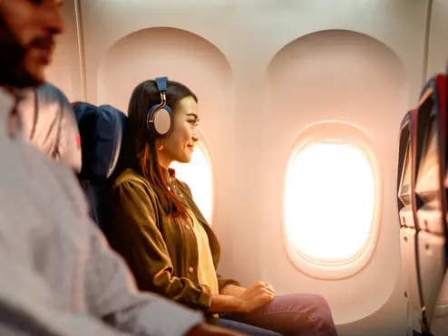 Delta customer on board an airplane wearing headphones sitting by a window, with warm sunlight illuminating the cabin and seatback screens visible.